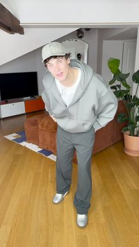 Gaiši pelēka džemperi ar rāvējslēdzēju, balta krekla, tumši pelēkas bikses un sudraba sporta apavi. Fonā redzams brūns dīvāns un lapots augs.