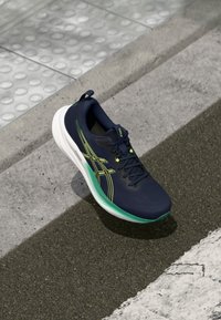 Marineblauw sportshoe met een gestructureerde bovenkant, groene accenten en een witte zool; heeft een verstevigde teen en een trekhaak aan de hiel.