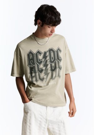 Junger Mann mit dunklem Haar trägt ein beiges AC/DC Band-T-Shirt und weiße strukturierte Hosen, schaut nach unten und hat eine Hand in der Tasche.