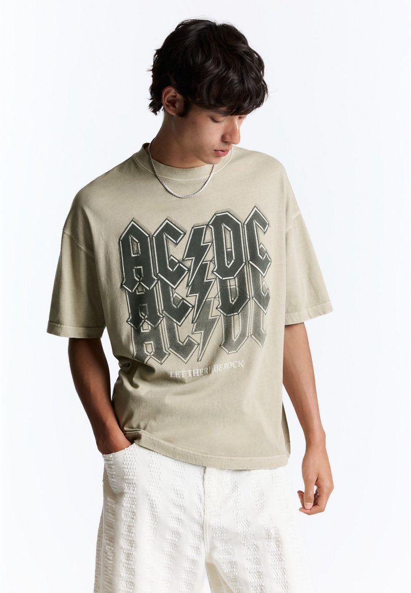 Jeune homme aux cheveux foncés portant un t-shirt beige du groupe AC/DC et un pantalon blanc texturé, regardant vers le bas avec une main dans la poche.