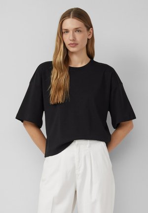 Donna con capelli biondi lunghi che indossa una maglietta nera a maniche corte larga infilata in pantaloni bianchi a vita alta su sfondo grigio.