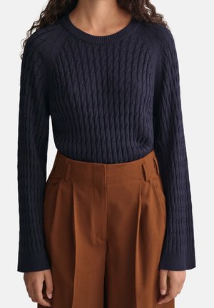 Pull en maille bleu marine avec un motif en câbles, col rond et manches longues, associé à un pantalon marron plissé taille haute.