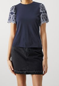 Kvinna som bär en marinblå topp med vita broderade korta ärmar och en svart denim minikjol med fransar.