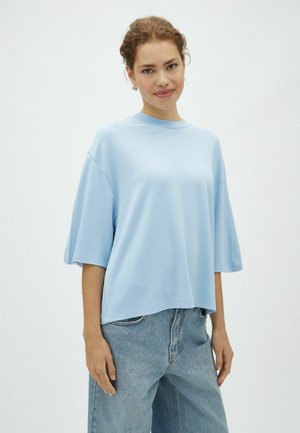Mladá žena s kučeravými vlasmi, ktorá má na sebe svetlomodrú oversized krátkym rukávom košeľu a voľné džínsové nohavice, stojí na jednoduchom pozadí.
