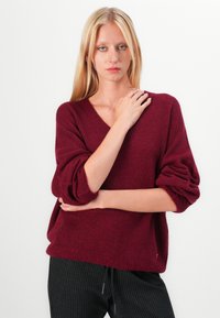 Maglione a maglia borgogna con scollo a V, maniche ampie e vestibilità comoda, abbinato a pantaloni neri a righe sottili.