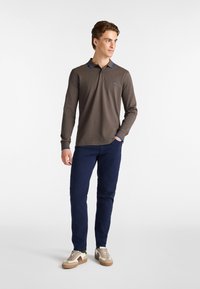 Polo a maniche lunghe marrone con colletto e logo blu, abbinato a pantaloni blu navy e scarpe da ginnastica beige con dettagli marroni.