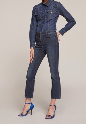 Kobieta w ciemnej jeansowej koszuli i dżinsach z niebieskimi sandałami na wysokim obcasie, stojąca z jedną ręką na biodrze na jednolitym tle.