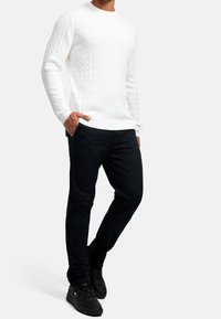 Weißer Pullover mit Zopfmuster und rundem Ausschnitt, kombiniert mit dunkler, schmal geschnittener Hose und schwarzen Sneakern, mit glatter Textur und gerippten Details.