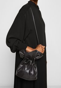 Černá kožená kabelka ve tvaru kbelíku s nařaseným vrškem, hladkým povrchem a úzkým ramenním popruhem, držená v ruce na jednobarevném pozadí.