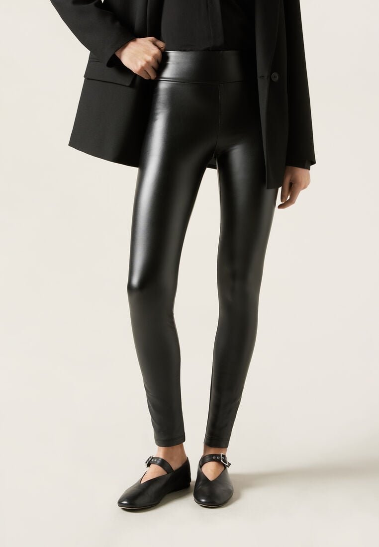 Person iført sorte leggings i kunstlæder, sort blazer og sorte Mary Jane-flade sko med ankelstropper foran en neutral baggrund.