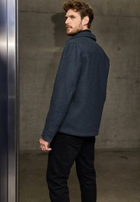 Kolfiberullblandad jacka med texturerad yta, knäppning vid ärmsluten och klassisk krage, kombinerad med svarta byxor mot en betongbakgrund.