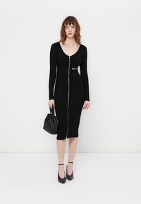 Vestido midi preto com nervuras, mangas longas e decote em V profundo, apresentando um fecho éclair frontal e uma silhueta ajustada, combinado com uma bolsa preta.