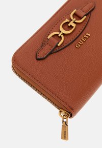 Portafoglio zip marrone realizzato in materiale sintetico testurizzato. Presenta un logo in tono oro e un accento a catena. Comprende una zip con un tirazip brandizzato.