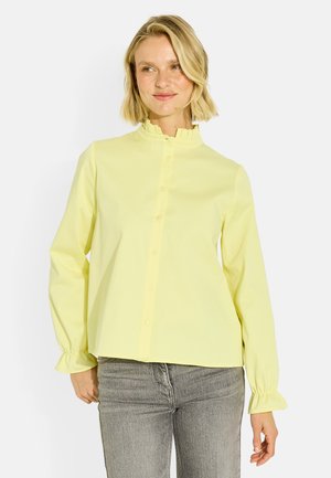 Femme blonde portant un chemisier jaune pâle à manches longues avec col et poignets volantés, associé à un jean gris, debout sur fond blanc.