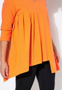 Leuchtend orangefarbene Tunika mit V-Ausschnitt, ausgestattet mit einem plissierten Design und drei Viertelärmeln, kombiniert mit schwarzen Leggings. Aus weichem Baumwollstoff.