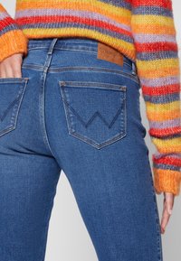 Blå denimjeans med høy midje, to baklommer med chevron-stikking og et skinnlignende logo-patch i midjen.