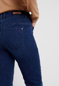 Persona vestida con jeans azul oscuro y un suéter marrón, con una mano metida en el bolsillo delantero contra un fondo liso.