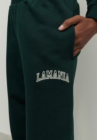 Žalios sportinės kelnės, pagamintos iš minkštos medžiagos, su kišene ir balta siuvinėta "LAMANIA" logotipu. Lygi tekstūra ir laisvas kirpimas.