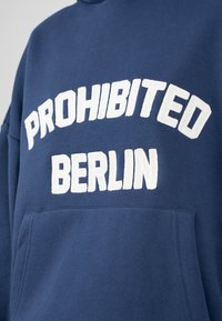 Sudadera azul marino con texto bordado en blanco "PROHIBITED BERLIN." Cuenta con un bolsillo tipo canguro y una tela texturizada. Diseño simple y casual.