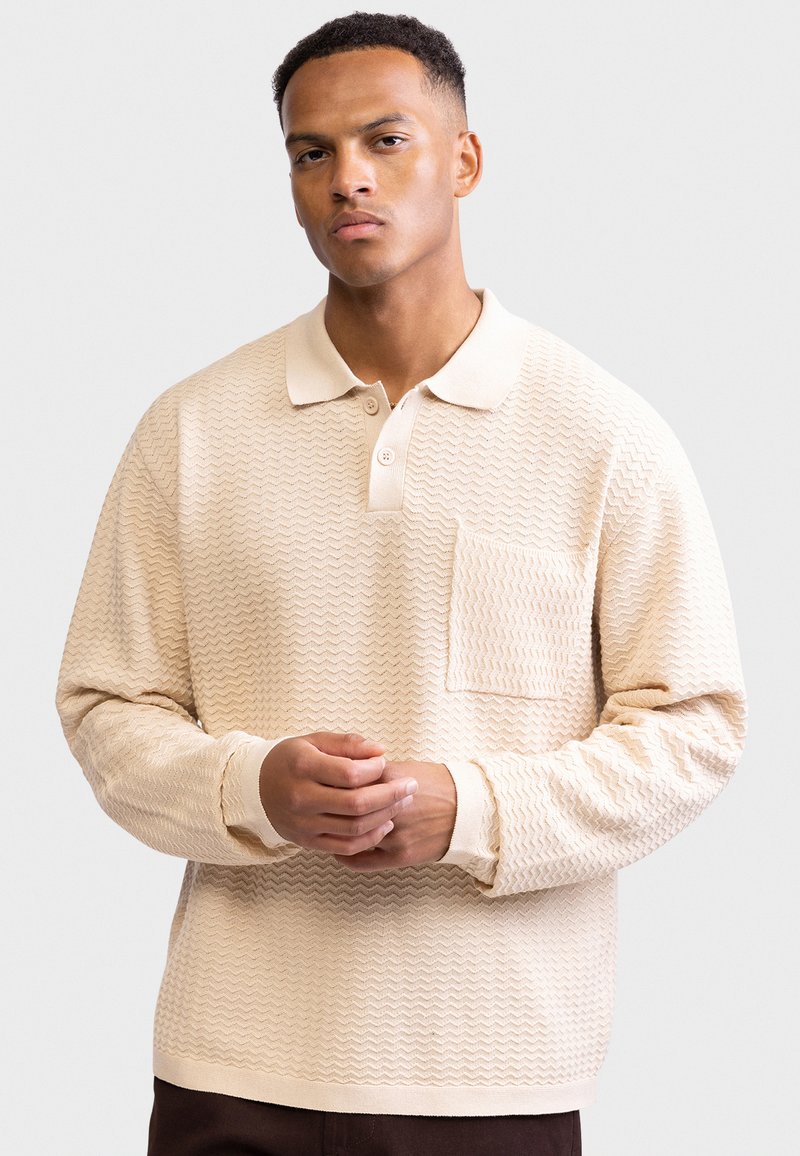 Beige textured polo shirt with a pocket, featuring a collar and button placket. Long sleeves with ribbed cuffs, made from a woven fabric.