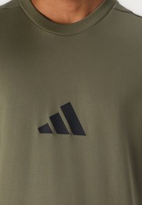 Oliven grønn bomulls T-skjorte med et marineblått Adidas-logo og tre diagonale striper. T-skjorten har rund halslinning og ribbestrikket kant.