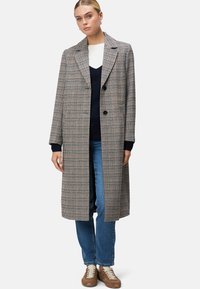 Lang gewatteerde jas met houndstooth-patroon in beige, zwart en bruin; heeft een notch-kraag, twee knopen en oversized zakken. Gekocht met een marineblauwe top en jeans.
