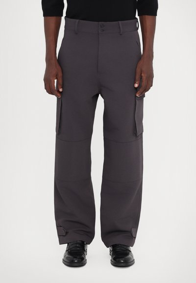 Pantalones de pierna ancha de color gris oscuro, confeccionados en un tejido suave, con bolsillos laterales, cierre de botón y puños ajustables. Combinados con zapatos negros brillantes.