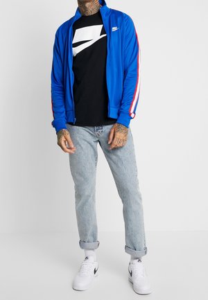 Blaue Sportjacke mit roten und weißen Streifen, schwarzes T-Shirt mit Logo, hellblaue Jeans und weiße Turnschuhe. Glatter Stoff, lockere Passform.