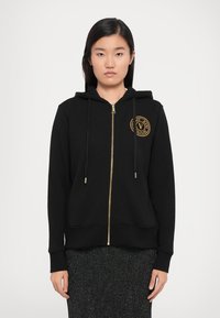 Black zip-up hoodie made of cotton blend fabric. Features a gold zipper, drawstrings, and a circular logo patch on the chest.