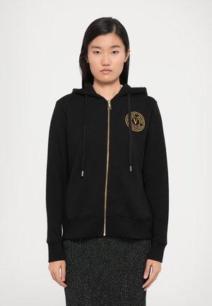 Black zip-up hoodie made of cotton blend fabric. Features a gold zipper, drawstrings, and a circular logo patch on the chest.