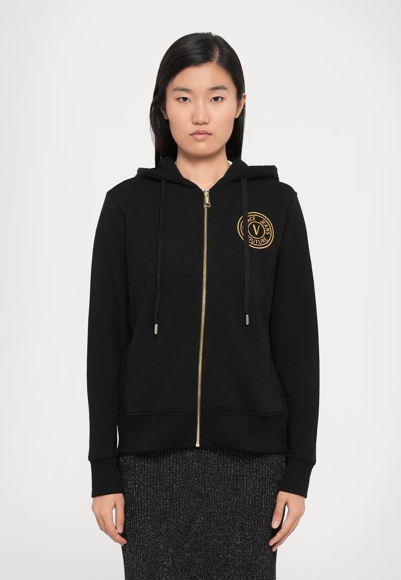Black zip-up hoodie made of cotton blend fabric. Features a gold zipper, drawstrings, and a circular logo patch on the chest.