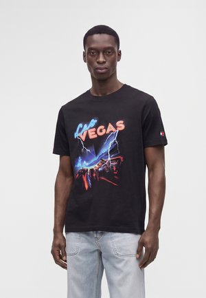 Άντρας με μαύρο T-shirt με νέον κείμενο "Las Vegas" και γραφικό αστραπής, σε συνδυασμό με ανοιχτό μπλε τζιν, στέκεται μπροστά σε απλό φόντο.