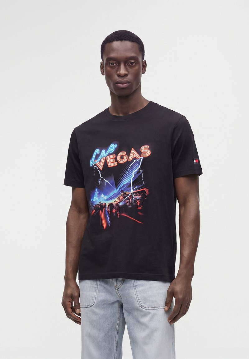 Άντρας με μαύρο T-shirt με νέον κείμενο "Las Vegas" και γραφικό αστραπής, σε συνδυασμό με ανοιχτό μπλε τζιν, στέκεται μπροστά σε απλό φόντο.