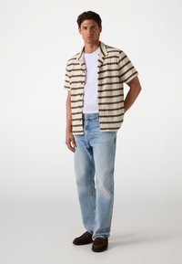 Camisa de manga corta a rayas en color crema y oliva, combinada con jeans de mezclilla azul claro y zapatos marrones sin cordones. Pose independiente contra un fondo claro.