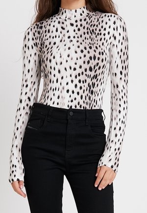 Femme portant un haut blanc à manches longues avec des taches noires et un pantalon noir taille haute sur un fond uni.