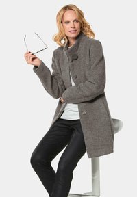 Gray wool coat with a high collar, featuring two large buttons and a relaxed fit. Model seated, holding glasses, wearing black pants.
