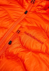 Giacca isolante arancione con texture trapuntata, dotata di una zip centrale e logo ricamato "PeakPerformance" di colore rosso a contrasto.