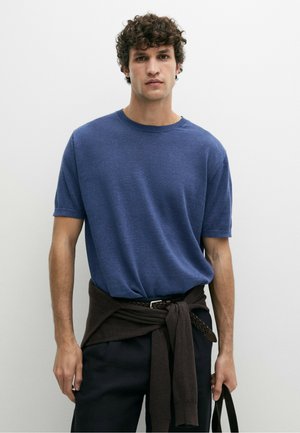 Mann mit lockigem Haar, der einen blauen kurzärmligen Pullover und schwarze Hose trägt, braunen Pullover um die Taille gebunden, hält einen schwarzen Taschenriemen.