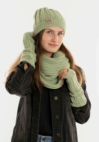 Lichtgroene gebreide beanie, sjaal en vingerloze handschoenen met ribbeltextuur. De sjaal is om de nek gewikkeld en toont een gelaagd ontwerp.