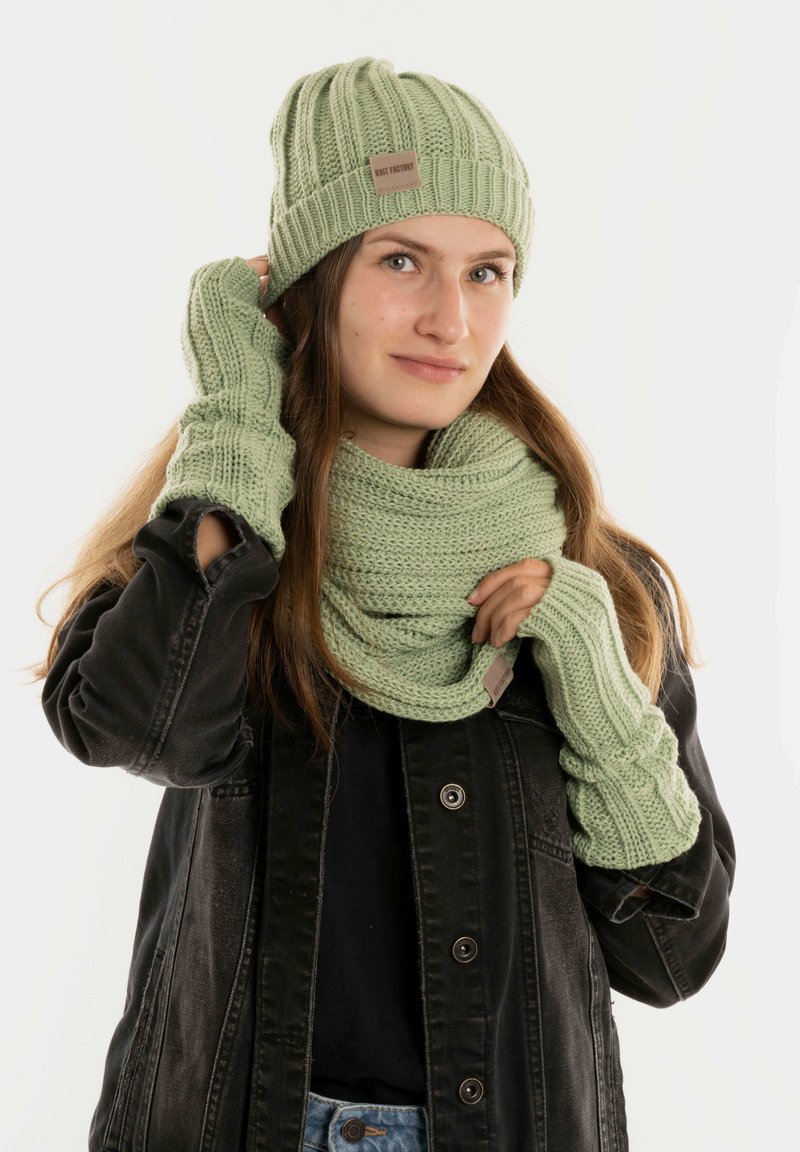 Lichtgroene gebreide beanie, sjaal en vingerloze handschoenen met ribbeltextuur. De sjaal is om de nek gewikkeld en toont een gelaagd ontwerp.