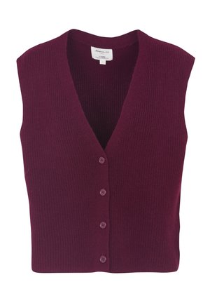 Femme portant un blazer beige à carreaux, une chemise rayée, un gilet bordeaux, un jean foncé avec revers, des chaussettes beiges et des mocassins foncés, debout devant un fond uni.