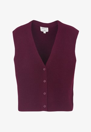 Vrouw draagt een beige geruit colbert, gestreept shirt, bordeauxrode bodywarmer, donkere jeans met omslag, beige sokken en donkere loafers, staand tegen een effen achtergrond.