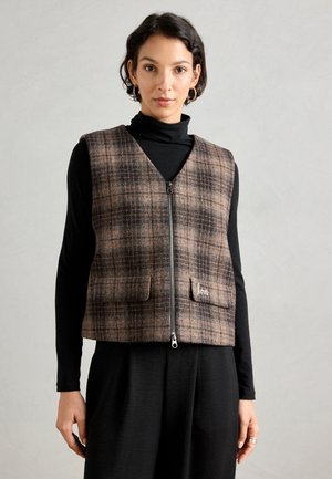 En kvinde iført en brunternet zip-up vest over en sort turtleneck og sorte bukser, står imod en ensfarvet grå baggrund.