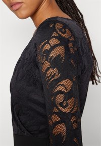 Μαύρη μπλούζα με υφή στο σώμα και δαντελωτά μανίκια με λουλουδάτο μοτίβο. Η δαντέλα είναι ημι-διάφανη με περίπλοκες σχεδίες.