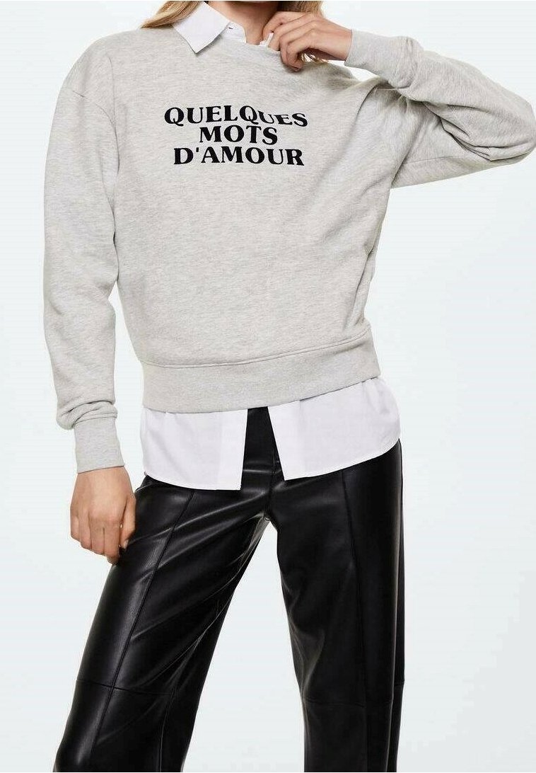 Felpa grigio chiaro con scritta nera "QUELQUES MOTS D'AMOUR", indossata sopra una camicia bianca con colletto e pantaloni di pelle neri.