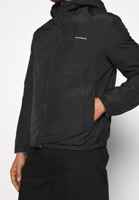 Svart lättviktsjacka med huva, som har dragkedjestängning och elastiska muddarna. Tyget är slätt med en subtil glans; liten logotyp på bröstet.