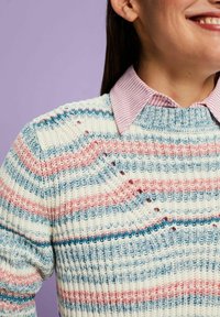 Personne souriante portant une chemise à col rayée rose sous un pull en tricot rayé bleu, rose et blanc sur un fond violet.
