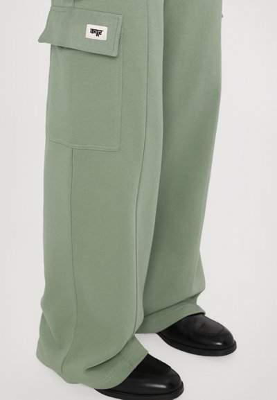 Light green sweatpants with a wide-leg design, featuring side pockets and black logo patch. Paired with black, smooth-textured shoes.