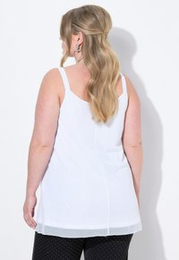 Top bianco senza maniche con un orlo inferiore leggermente trasparente, caratterizzato da una linea dritta e una texture liscia. Indossato con pantaloni neri a pois.