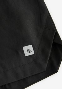 Black fabric garment corner featuring a grey, square logo with a triangular design. The fabric has a smooth, matte texture.